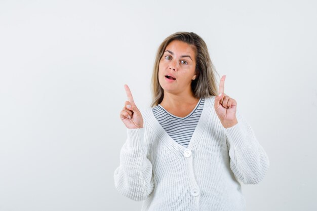 Fille blonde pointant vers le haut avec l'index en t-shirt rayé, cardigan blanc et pantalon en jean et à la recherche de bonheur. vue de face.