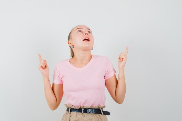 Fille blonde pointant vers le haut en t-shirt, pantalon et à la recherche d'espoir. vue de face.