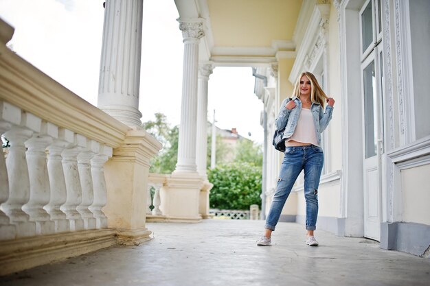 Une fille blonde porte un jean avec un sac à dos posé contre une maison vintage