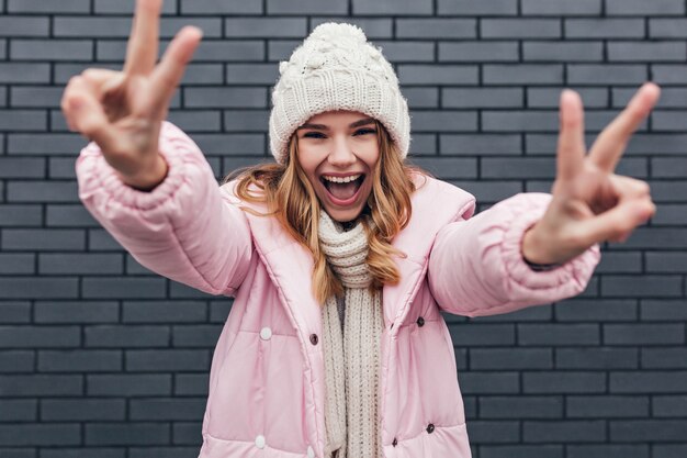 Fille blonde positive au chapeau exprimant des émotions heureuses. Dame caucasienne inspirée en vêtements d'hiver en riant