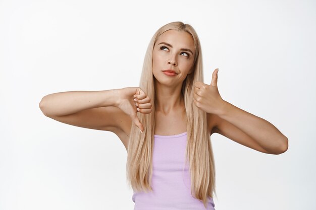 Une fille blonde réfléchie montre des pouces de haut en bas, prend une décision, pèse le pour et le contre, aime ou n'aime pas, se tient sur blanc