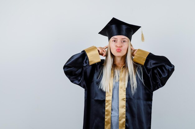 Fille blonde en robe de graduation et casquette bouchant les oreilles avec l'index, envoyant des baisers et ayant l'air mignon