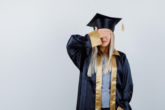 Fille blonde en robe de graduation et casquette couvrant les yeux avec la main et à la recherche de sérieux