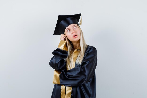 Fille blonde en robe de graduation et casquette debout dans une pose de réflexion et à la pensive