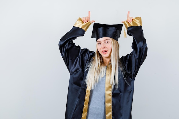 Fille blonde en robe de graduation et casquette pointant vers le haut avec l'index et à la mignonne