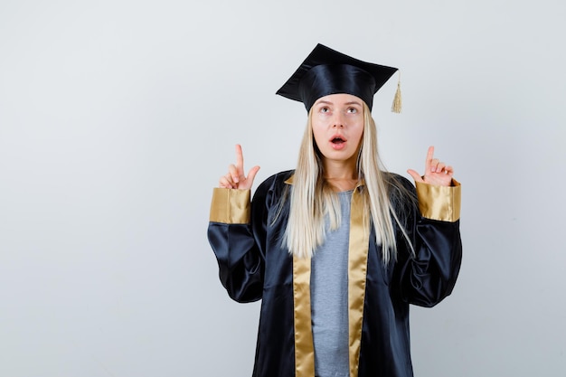 Fille blonde en robe de graduation et casquette pointant vers le haut avec l'index et à la mignonne