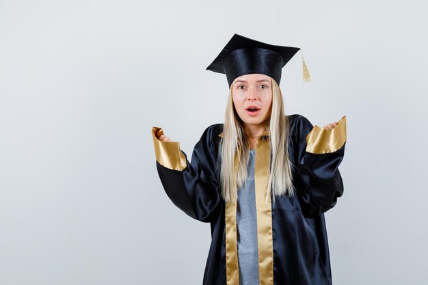 Fille Blonde S'étirant Les Mains De Manière Interrogative En Robe De Graduation Et Casquette Et L'air Surpris.