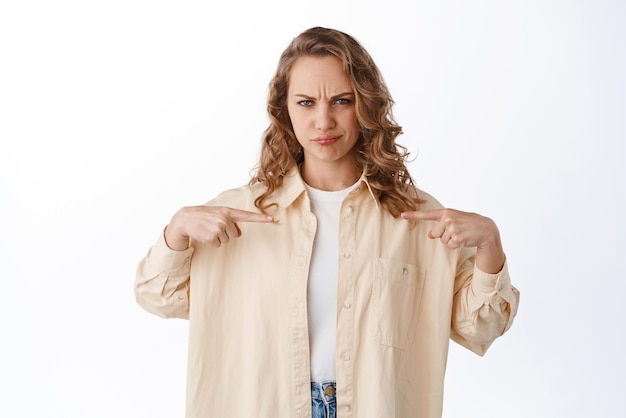 Fille Blonde Sceptique Et Douteuse Grimaçant En Se Pointant Du Doigt Avec Une Expression De Visage Malheureux Dérangé Debout Sur Fond Blanc