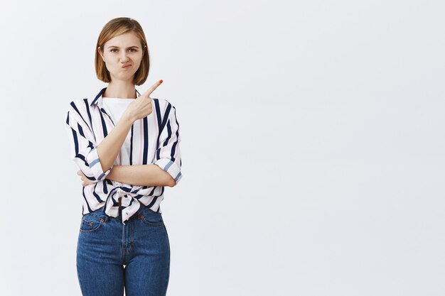 Fille blonde sceptique et mécontente souriant, pointant le coin supérieur droit