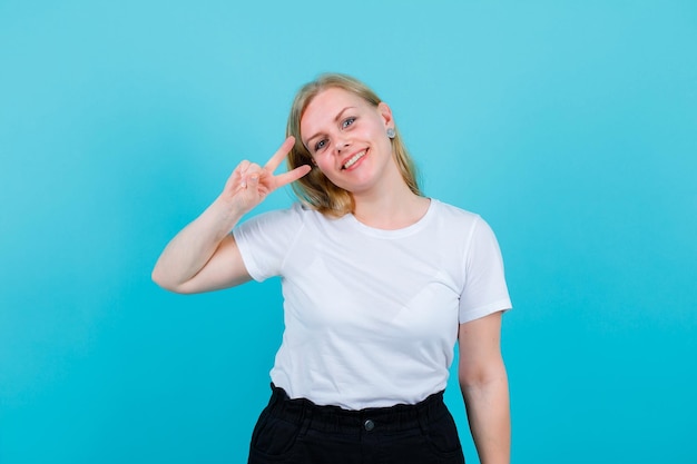 Une fille blonde souriante montre deux gestes en tenant la main près de l'œil sur fond bleu