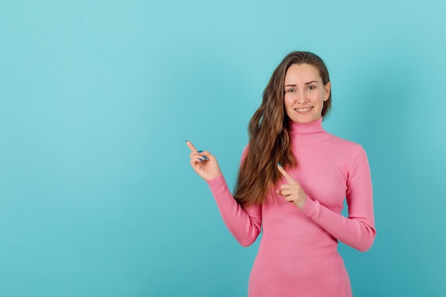 Une fille blonde souriante pointe vers la gauche avec des index sur fond bleu