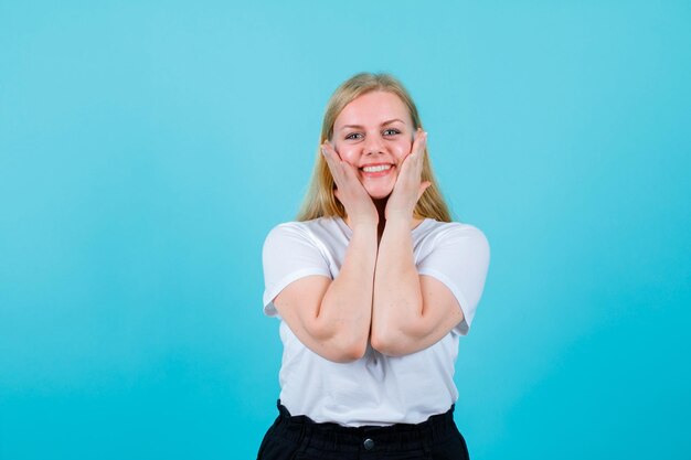 Une fille blonde souriante regarde la caméra en mettant les mains sur les joues sur fond bleu