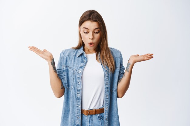 Fille blonde surprise regardant vers le bas avec les mains écartées, regardant l'espace de copie en bas étonné, debout dans une chemise en jean et un t-shirt blanc contre le mur du studio