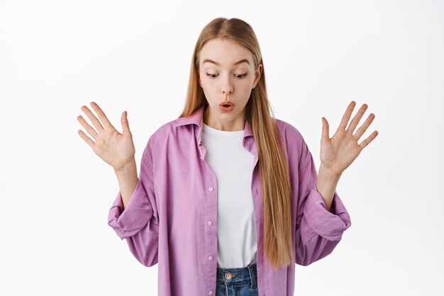 Une fille blonde surprise regarde vers le bas et dit wow, écarte les mains sur le côté étonné, laisse tomber quelque chose, regarde le logo de la bannière ci-dessous, debout contre le mur blanc