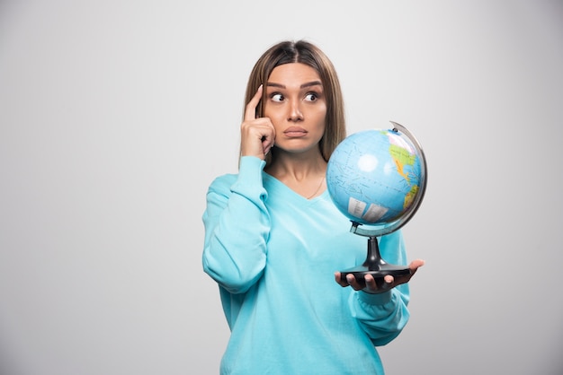 Fille blonde en sweat-shirt bleu tenant un globe, réfléchissant attentivement et essayant de se souvenir.