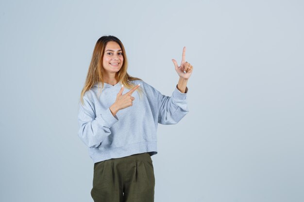 Fille blonde en sweat-shirt bleu vert olive et pantalon pointant vers le haut et à la radieuse, vue de face.