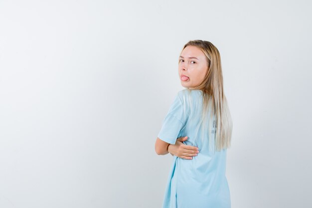 Fille blonde en t-shirt bleu tournant en arrière, collant la langue tout en posant et à la jolie vue de face.