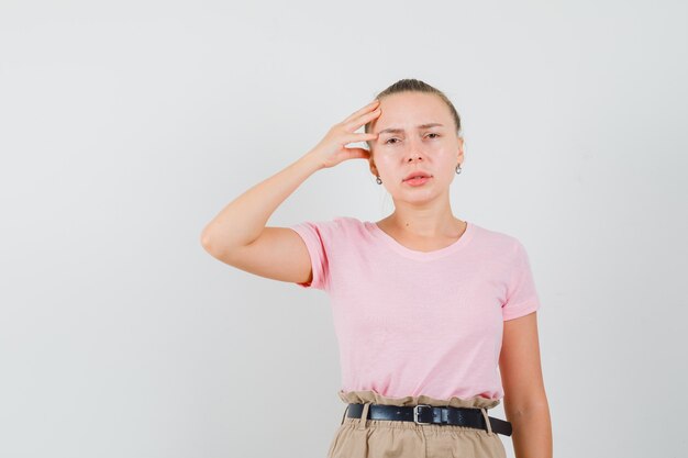 Fille blonde en t-shirt, pantalon ayant de forts maux de tête et regardant triste, vue de face.