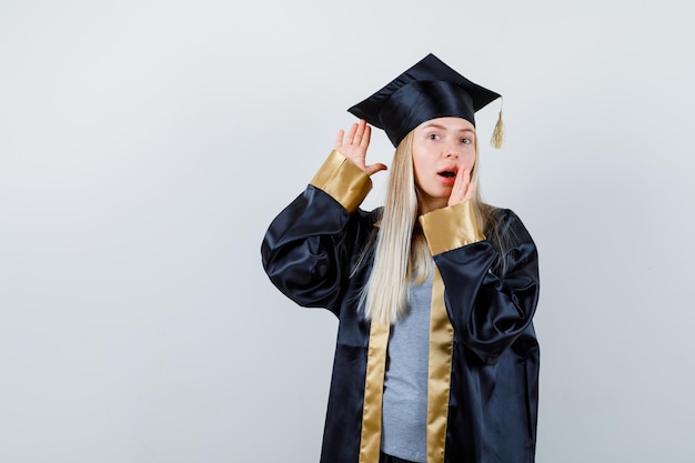 Photo gratuite fille blonde tenant la main près de l'oreille pour entendre quelque chose, tenant la main près de la bouche en robe de graduation