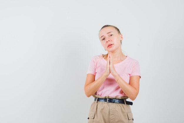 Fille blonde tenant par la main en signe de prière en t-shirt, pantalon et à la recherche d'humble. vue de face.