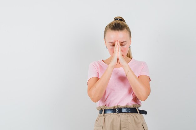 Fille blonde tenant par la main en signe de prière en t-shirt, pantalon et à la vue calme, de face.