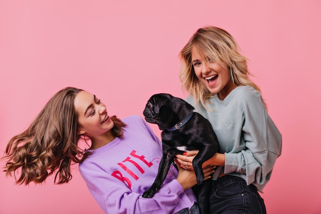 fille blonde tenant son chien et riant. Portrait intérieur de dames raffinées s'amusant ensemble et jouant avec un chiot.