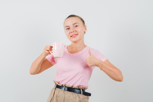 Fille blonde tenant une tasse de boisson, montrant le pouce vers le haut en t-shirt, pantalon et à la joyeuse. vue de face.