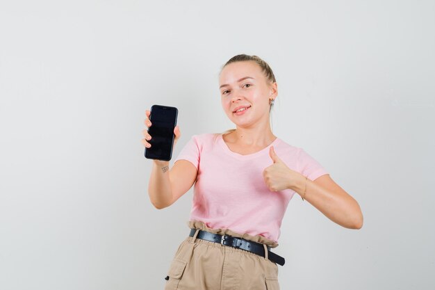 Fille blonde tenant un téléphone portable, montrant le pouce vers le haut en t-shirt, pantalon et à la joyeuse vue de face.