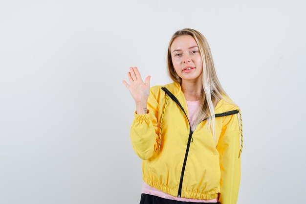 Fille blonde en veste jaune, agitant la main pour saluer et avoir l'air attrayant