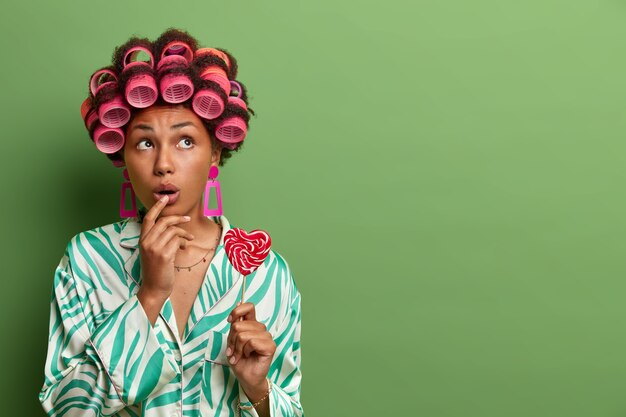 Une fille de bonbons surprise réfléchie regarde ci-dessus, pose avec une délicieuse sucette, porte des bigoudis pour faire des bigoudis parfaits, porte un pyjama, se tient contre un mur vert, un espace vide à droite