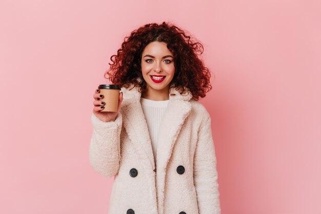 Fille bouclée aux yeux bleus avec rouge à lèvres vêtue d'un manteau de fourrure blanc écologique souriant et tenant un verre de café sur l'espace rose.