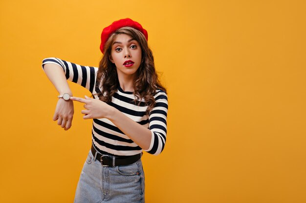 Fille bouclée en béret pointant vers sa montre. Jeune femme moderne aux cheveux ondulés en pull rayé posant sur fond isolé.