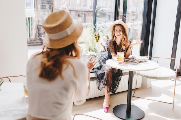 Fille bouclée en chemise blanche et chapeau racontant une histoire drôle à un ami aux cheveux longs, boire un cocktail