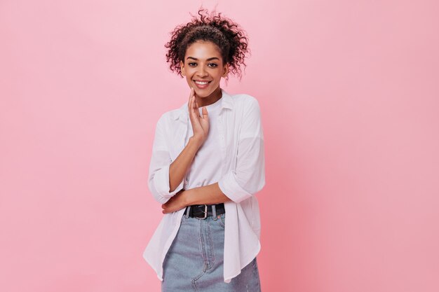 Une fille bouclée en chemise blanche sourit sur un mur rose