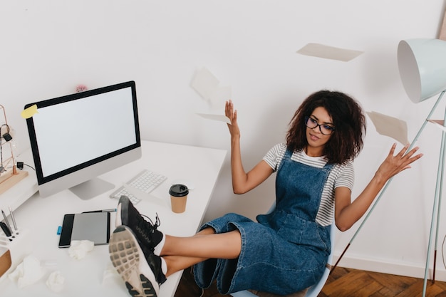 Photo gratuite fille bouclée noire disperse les documents assis sur une chaise avec les jambes sur la table