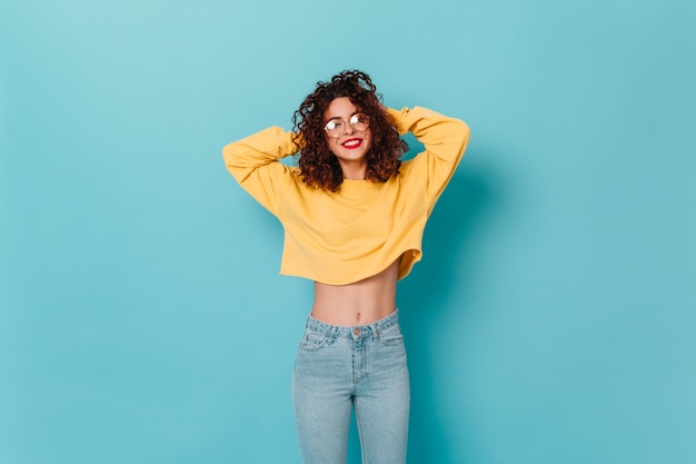 Fille bouclée avec rouge à lèvres sourit sincèrement. Femme portant des lunettes, un sweat-shirt court jaune et un jean posant sur l'espace bleu avec les bras levés.