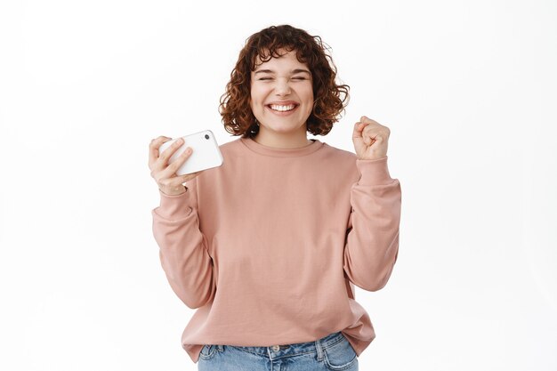 Une fille bouclée satisfaite célèbre, dit oui, atteint l'objectif, tient un smartphone horizontal et fait une pompe à poing, gagnant sur l'application de jeu vidéo, debout sur blanc.