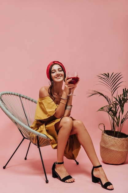 Fille bouclée souriante avec une robe élégante à bandeau lumineux et des chaussures sombres tenant un verre avec du vin rouge et assis sur une chaise cool
