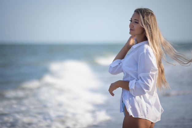 Une fille bronzée aux longs cheveux blonds dans un maillot de bain bleuté délicat et une chemise légère blanche, passe dans ses longs cheveux au bord d'une mer orageuse
