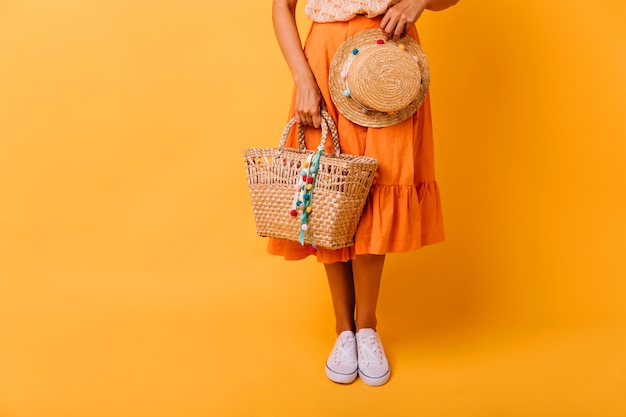 Fille bronzée en jupe orange et chaussures blanches debout sur jaune. Modèle féminin spectaculaire avec chapeau à la mode qui pose en studio.