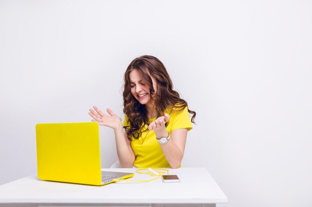 Une fille brune aux cheveux bouclés rit devant un ordinateur portable dans un étui jaune. Elle est assise derrière une table blanche et porte un t-shirt jaune. Il y a un smartphone sur un chargeur jaune sur la table.