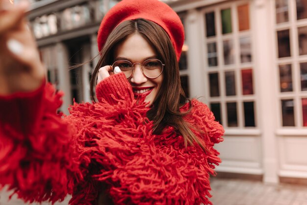 Fille brune aux cheveux longs avec manucure blanche et rouge à lèvres rouge fait selfie Photo d'une charmante femme à lunettes et tenue de laine rouge sur fond de maison