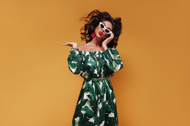 Fille brune bouclée inhabituelle avec des lunettes de soleil blanches et des boucles d'oreilles rouges dans une tenue cool à la mode soufflant des baisers et regardant dans l'appareil photo