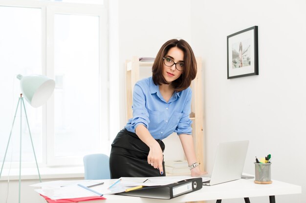 Fille brune en chemise bleue et jupe noire est debout près de la table au bureau. Elle posa sa main sur la table et la montra aux papiers. Elle regarde sérieusement la caméra.