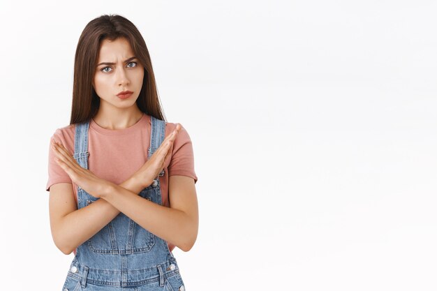 Fille brune en colère à l'air sérieux en salopette en jean, t-shirt, faisant arrêter quelqu'un par le haut, empêcher une mauvaise décision, secouer la tête en réponse négative, être en désaccord, interdire ou refuser l'offre