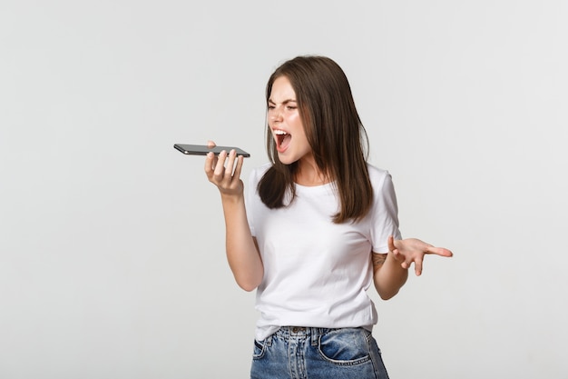 Fille brune en colère et énervée se disputant, enregistrer un message vocal sur un téléphone portable, blanc.