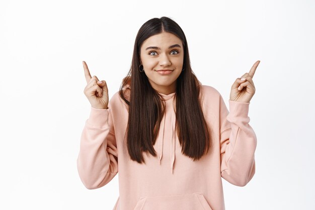 Fille brune confuse se sentant mal à l'aise, souriante et pointant les doigts vers le haut, montrant une publicité, debout contre un mur blanc