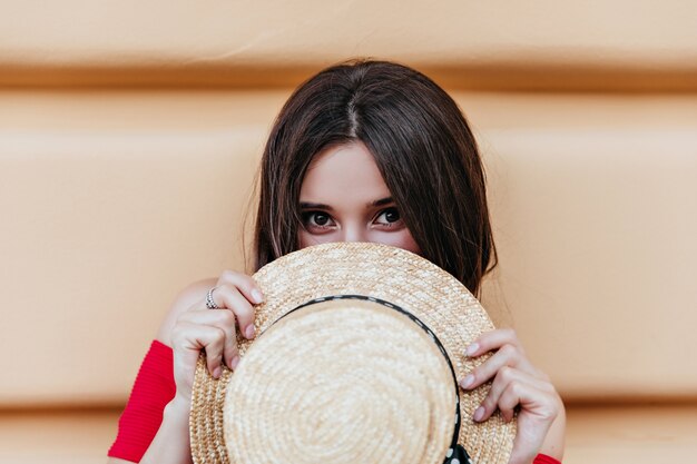 Fille brune drôle aux yeux sombres posant avec un chapeau de paille dans la rue. Dame sensuelle aux cheveux bruns exprimant des émotions positives.