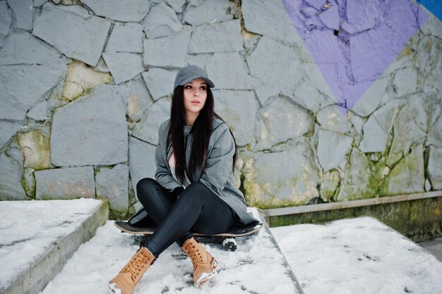 Fille brune élégante en casquette grise style de rue décontracté avec planche à roulettes le jour d'hiver contre un mur coloré