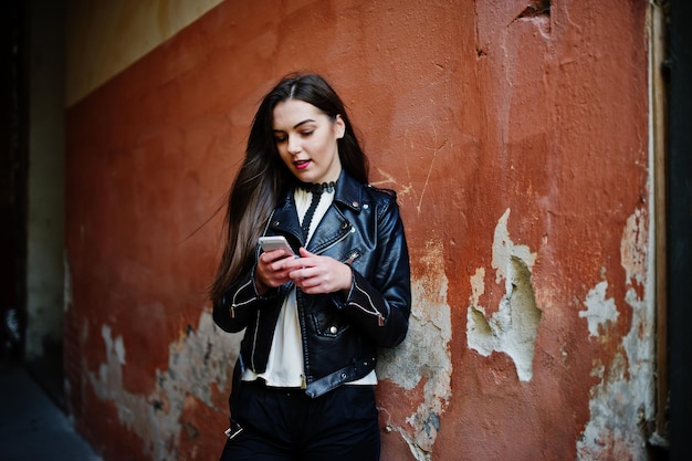 Une fille brune élégante porte une veste en cuir et un short avec un téléphone portable à la main contre le mur orange